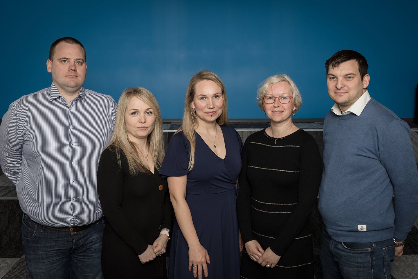 Group photo of five people standing together indoors against a blue wall.