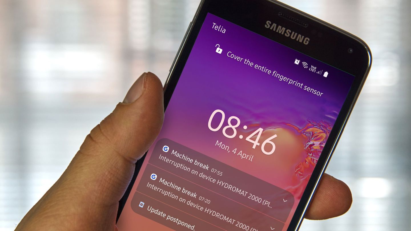 Close-up of a Samsung smartphone screen displaying notifications, including machine breaks and an update postponement, with a blurred window background.
