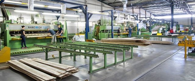 Workers operating machinery in a large wood manufacturing factory, with stacks of cut wooden planks and equipment for processing wood.