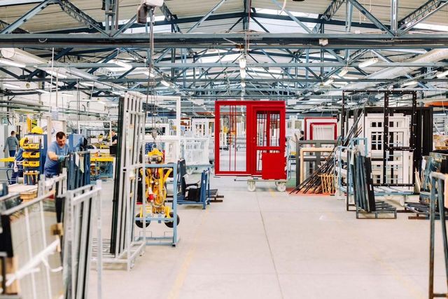 Manufacturing facility with metal frames, mostly white, with various window and door frames, some red and black, on mobile carts and units. Workers are assembling or inspecting parts, with high ceiling and exposed metal beams.